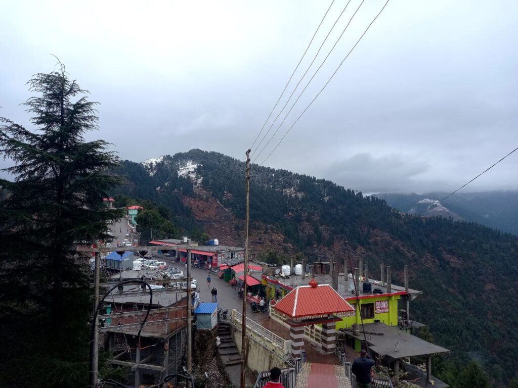 Village with colorful buildings on hilly terrain, snow-capped peaks, trees, and cloudy sky.