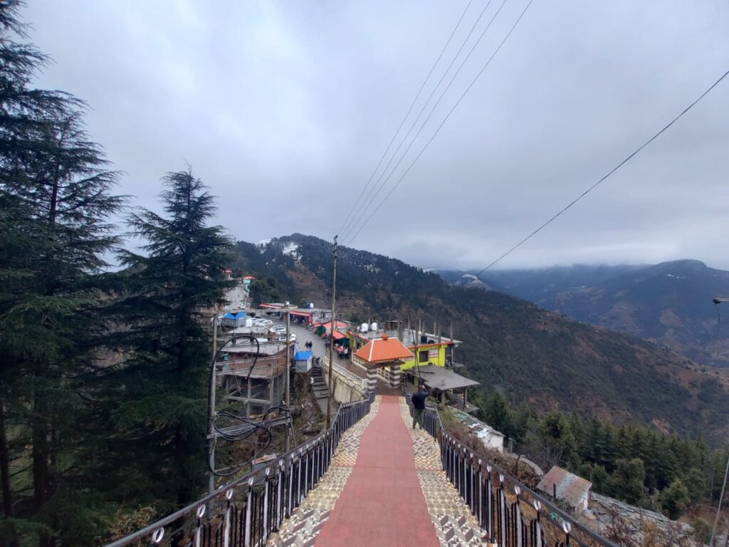 Pathway with railing leads to hilly village amid pine trees and mountains under cloudy sky.