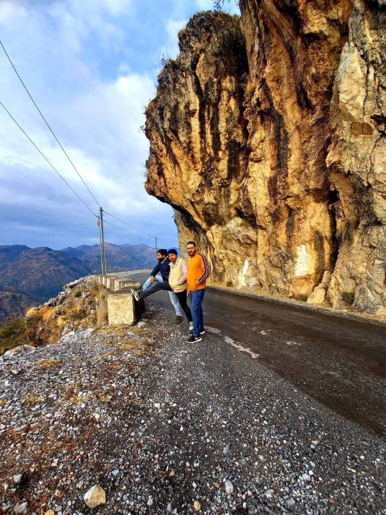 Three people stand by a curved mountain road with rocky cliffs and distant hills.