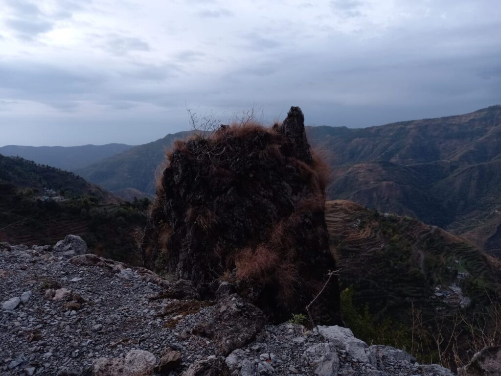 Rocky outcrop with sparse grass on hillside, overlooking mountains with terraced fields, overcast sky.