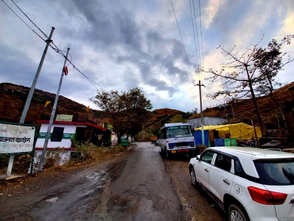 Rural road with bus, white car, buildings, colorful tarps, power lines, hills, cloudy sky.